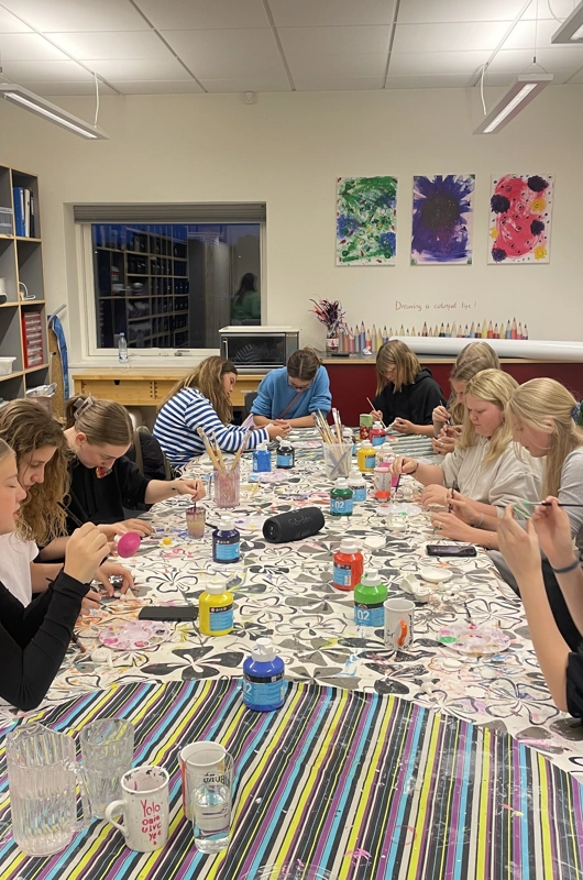 12 teenagepiger sidder rundt om et bord og maler