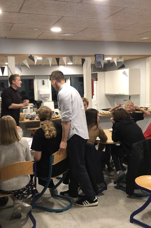 mange elever i et fælleslokale, sidder ved borde. Lærer står op.